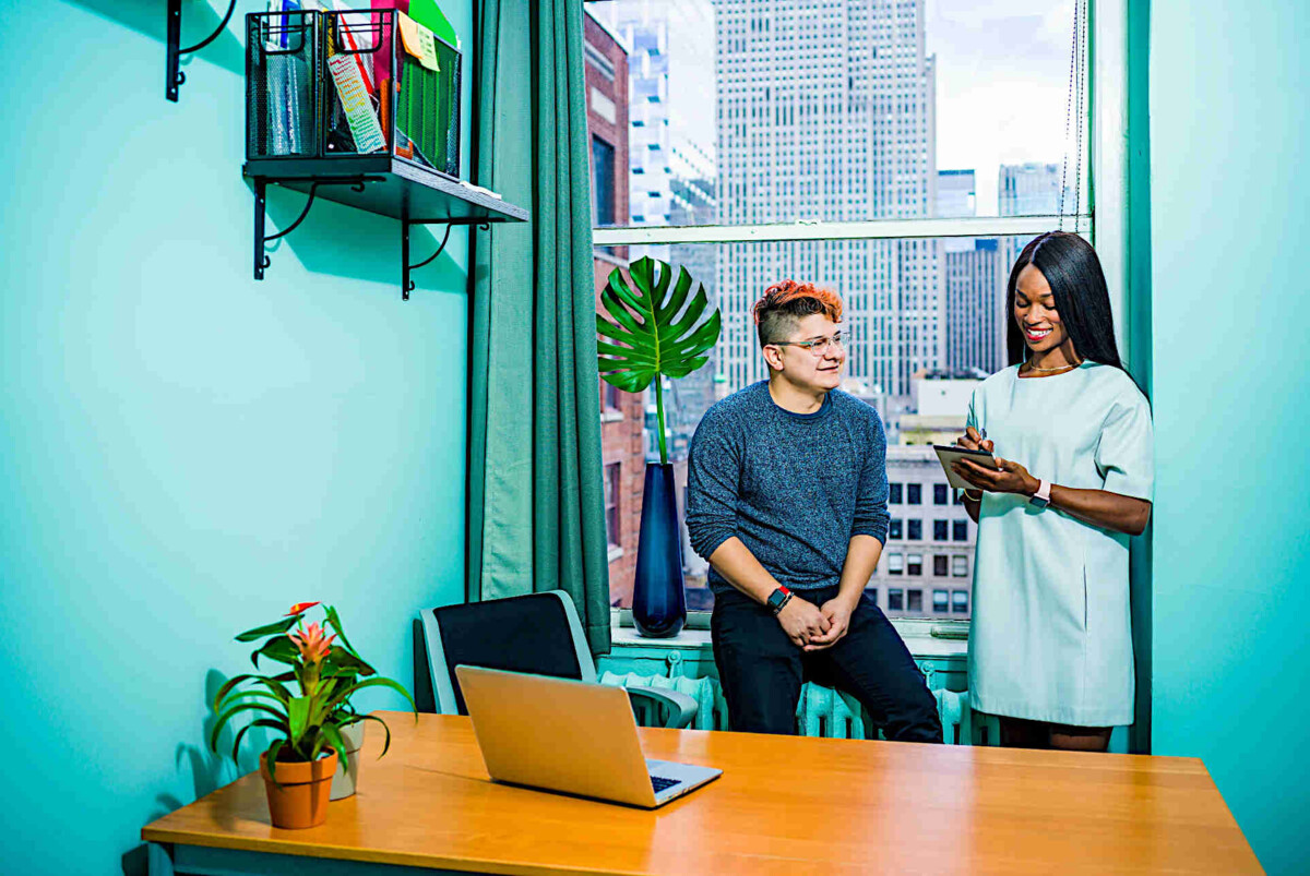 Zwei Kollegen wie vor einem Küchenfenster mit blauer Tapete. Er hat gefärbte Haare und sitzt sie ist farbig und steht. Auf dem Tisch stehen zwei Laptops.