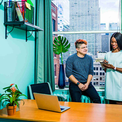 Zwei Kollegen wie vor einem Küchenfenster mit blauer Tapete. Er hat gefärbte Haare und sitzt sie ist farbig und steht. Auf dem Tisch stehen zwei Laptops.